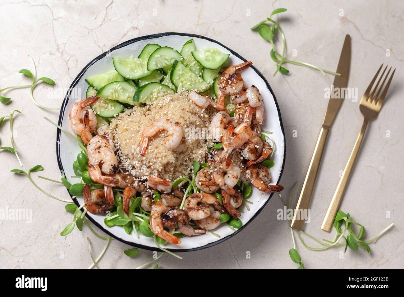 Quinoa with grilled prawns or shrimps, cucumber and micro greens on the marble background near golden folk and knife. Stock Photo