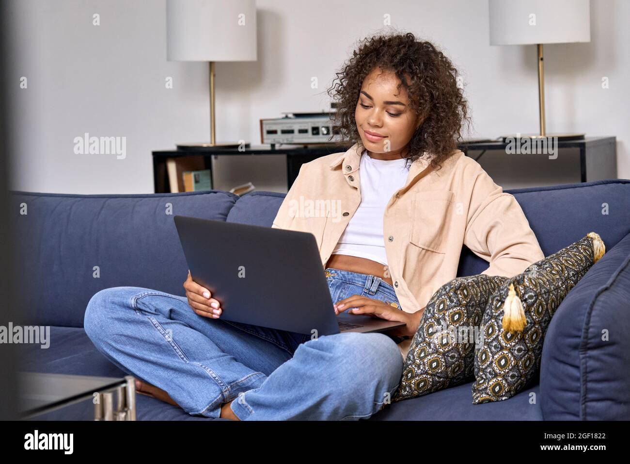 Happy young African woman using laptop computer sitting on sofa at home. Stock Photo