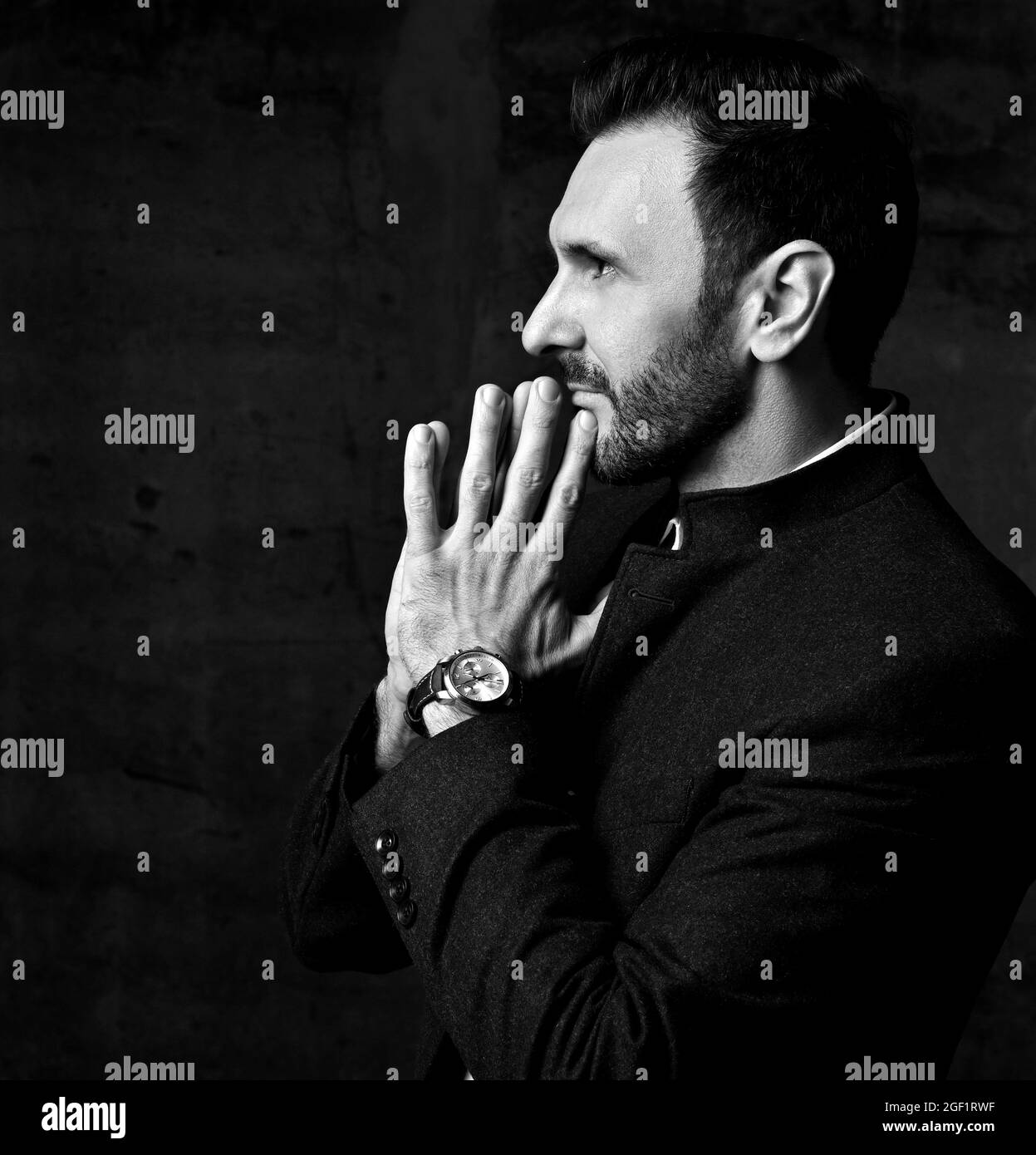 Black and white. Adult bearded man in jacket with mandarin collar stands sideways with hands put together at his lips Stock Photo