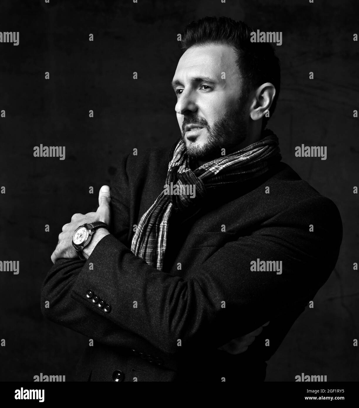 Black and white. Elegant bearded man in jacket and stylish scarf stands sideways holding arms crossed at chest Stock Photo