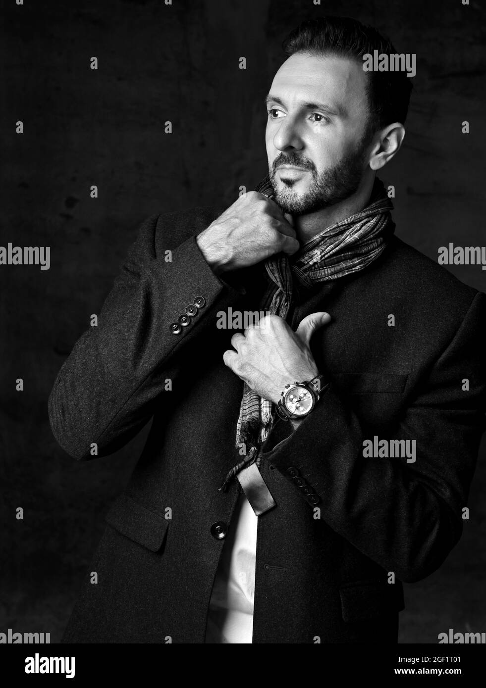 Black and white. Elegant unshaved adult man in jacket stands putting on his stylish scarf and looks aside Stock Photo