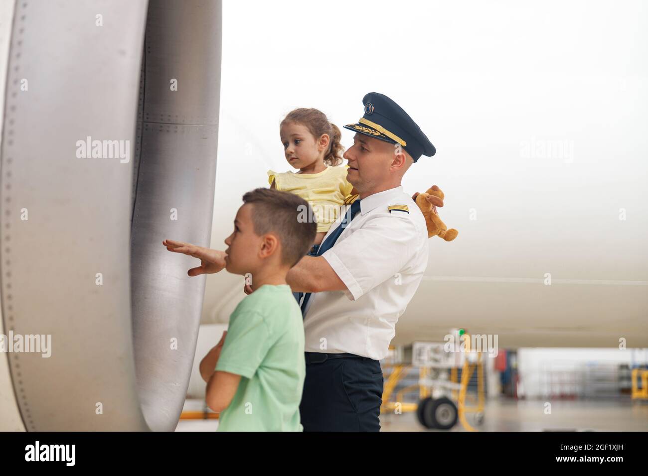 Pilot in uniform showing parts of airplane to two little kids who came to excursion at the aircraft hangar Stock Photo