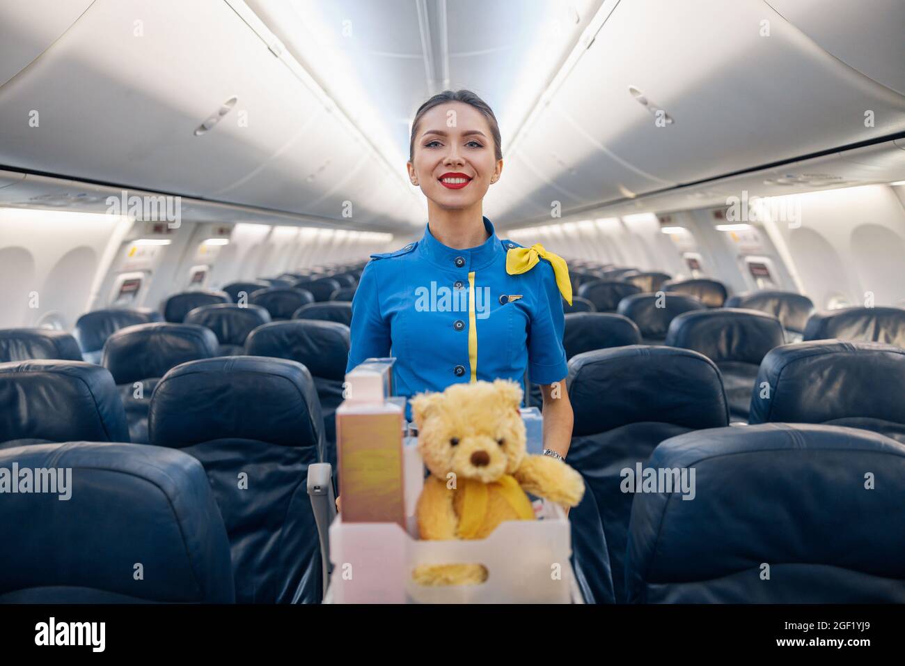 Attractive female air hostess in bright blue uniform smiling at camera while leading trolley cart with gifts, souvenirs through empty plane aisle Stock Photo