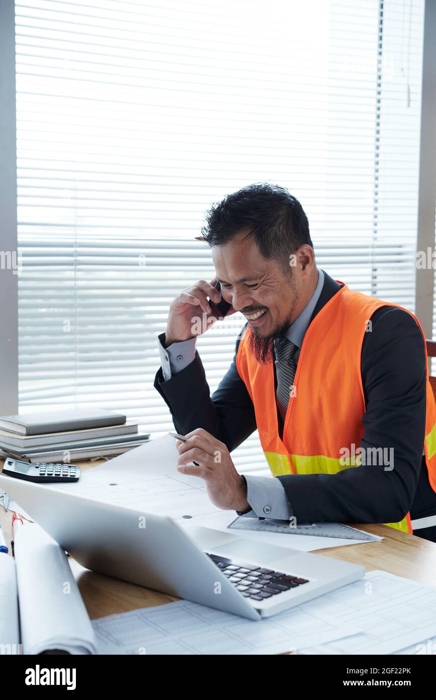Positive construction engineer working at office desk, checking blueprints and discussing readiness of construction site with contractor on phone Stock Photo