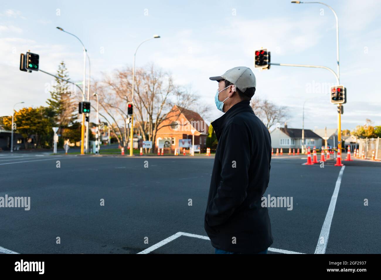 Man wearing a facemask waiting at the intersection for the green pedestrian light. Roadwork traffic cones along the street. Stock Photo