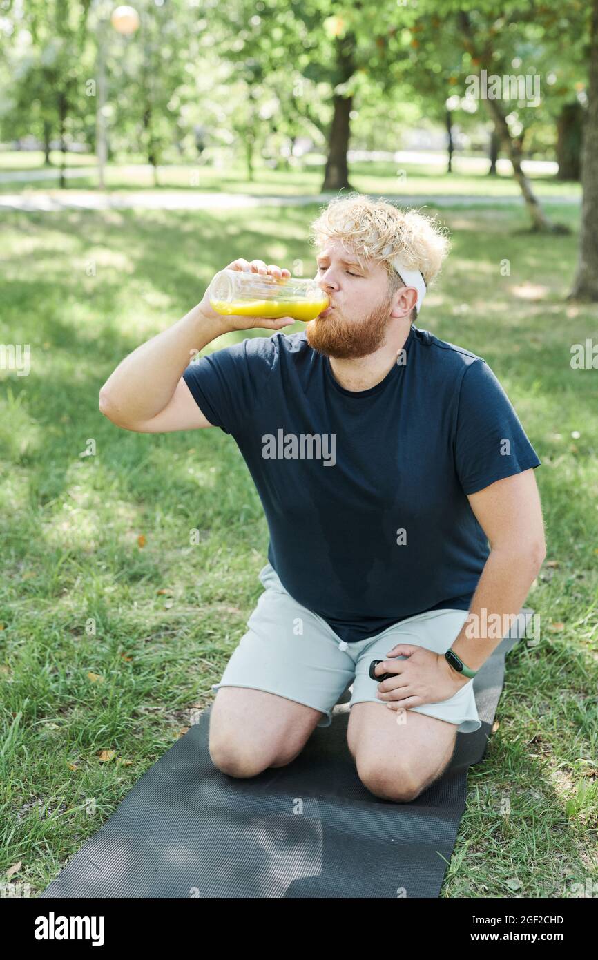 Overweight man sitting on exercise mat and drinking fresh juice during sports training in the