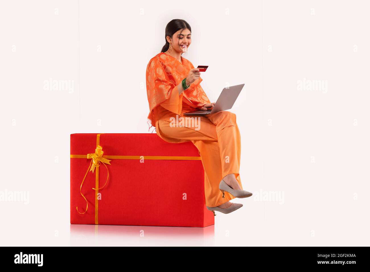 A young woman sitting on gift shaped stool making online transaction with credit card and laptop. Stock Photo