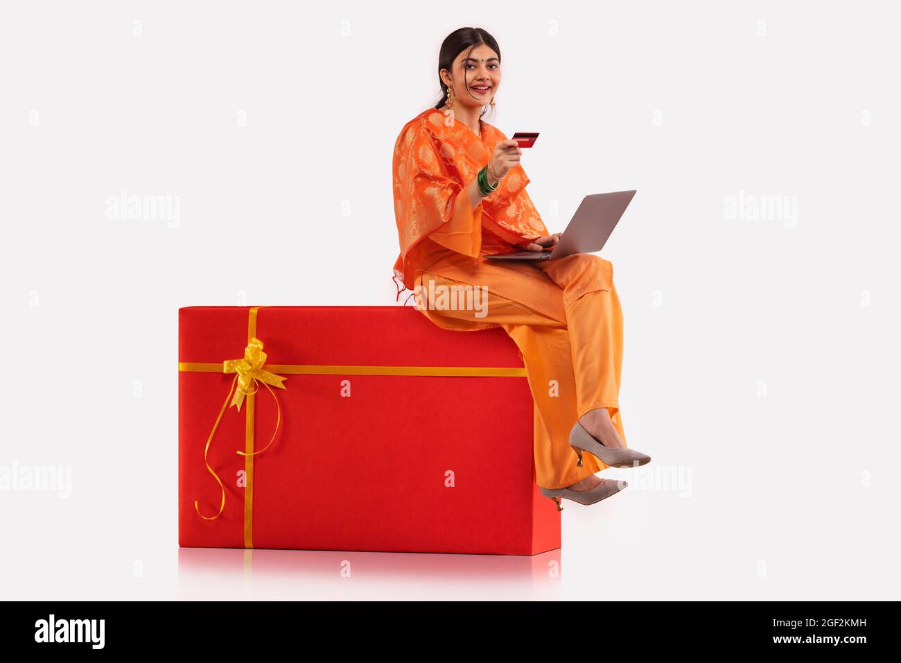 A young woman sitting on gift shaped stool making online transaction with credit card and laptop. Stock Photo