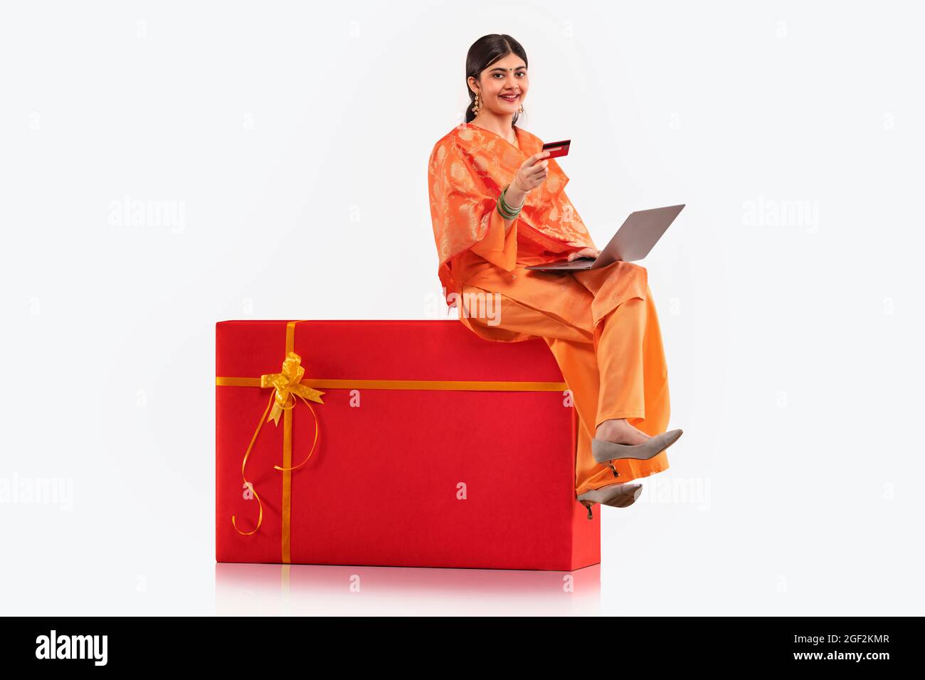 A young woman sitting on gift shaped stool making online transaction with credit card and laptop. Stock Photo