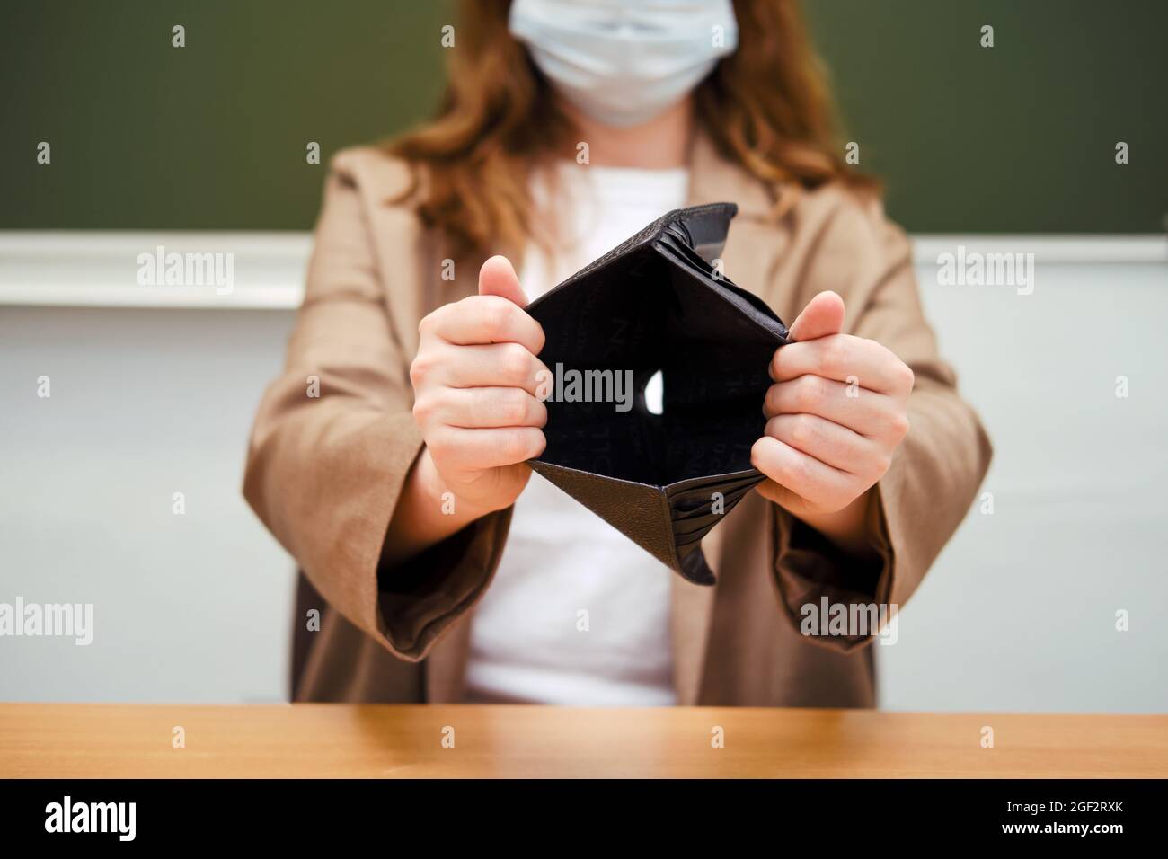 School teacher in a medical mask sits at a desk with an empty wallet ...