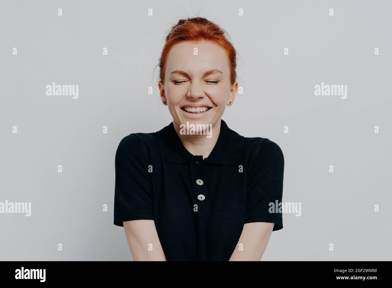Happy pleased red haired woman with closed eyes and toothy smile posing against grey wall Stock Photo