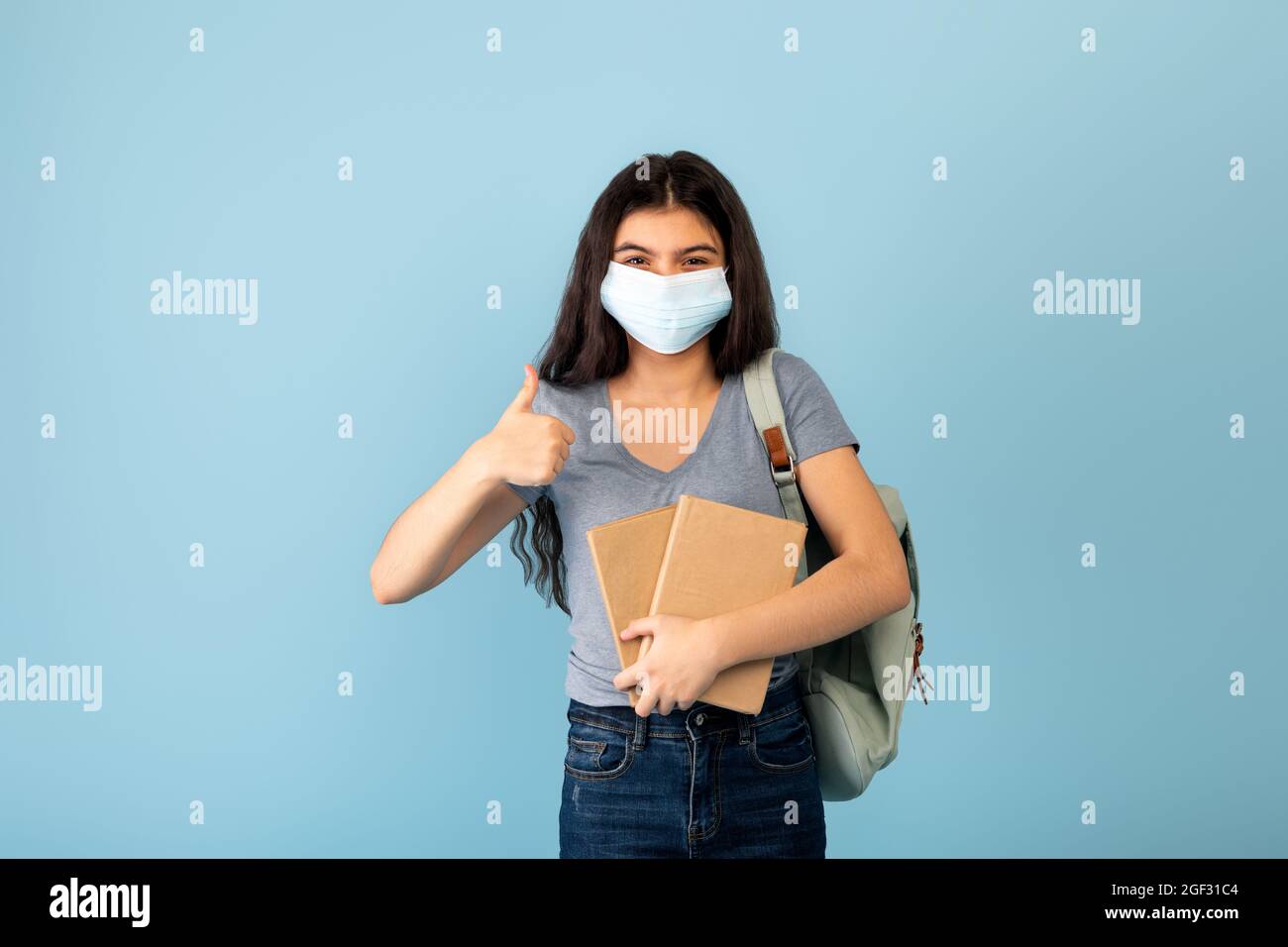 Funky Indian teen student in face mask with backpack holding books and showing thumb up gesture on blue background Stock Photo