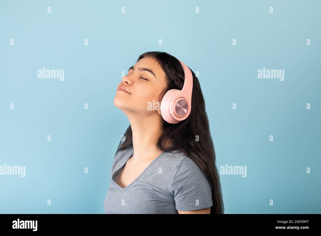 Attractive Indian teen girl enjoying her new stereo headset, listening to music with closed eyes over blue background Stock Photo