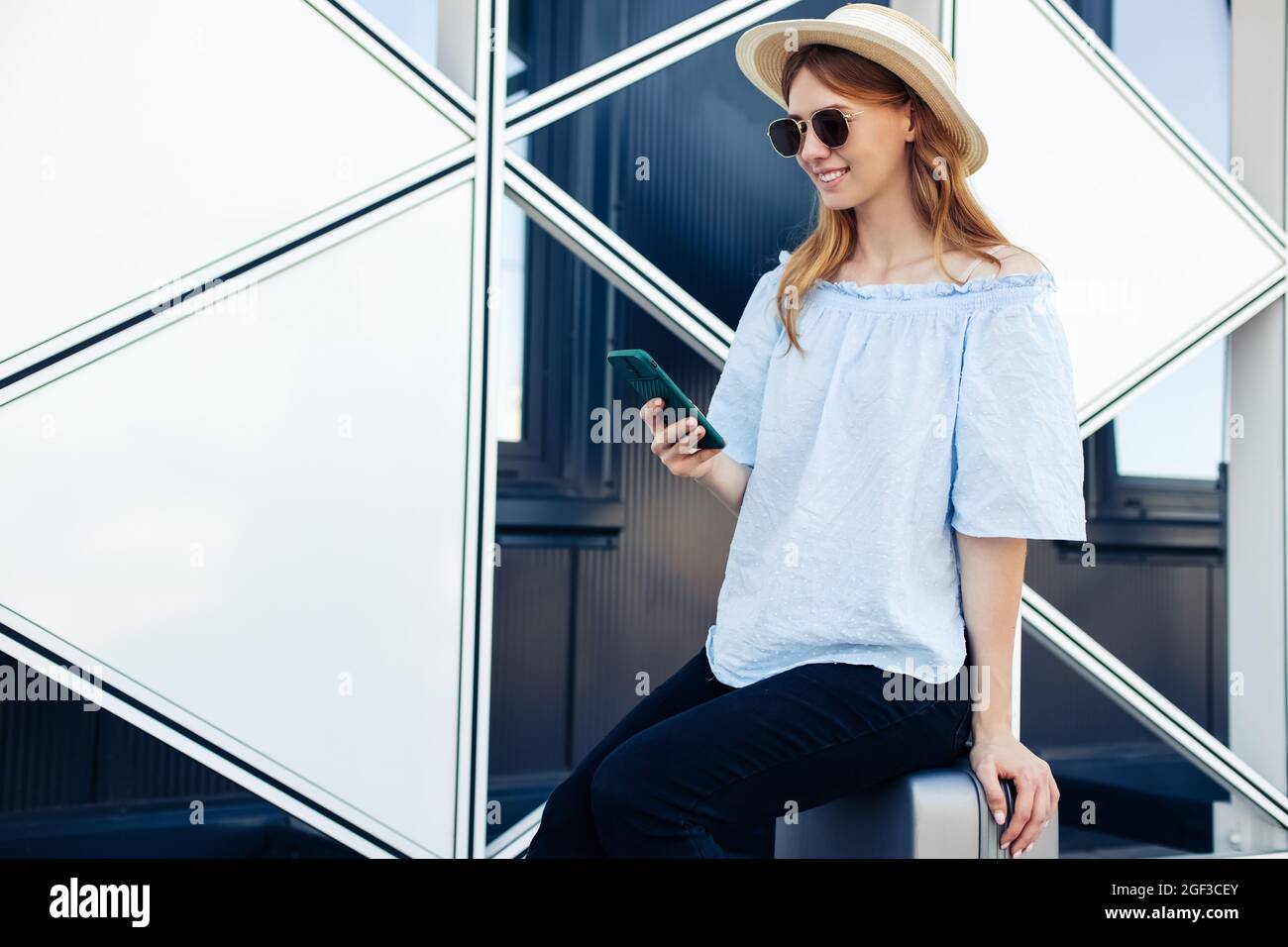 Smiling young woman dressed in summer clothes and hat sits on a suitcase near the airport, a traveler uses a mobile phone, Woman with luggage before t Stock Photo