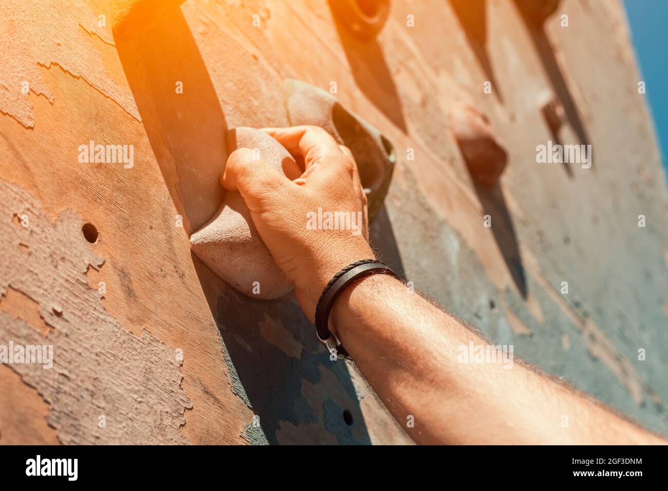 Close-up photo of male hand gripping climbing holds on worn wall outside. Man doing climbing exercise on artificial rock climbing wall at park. Stock Photo