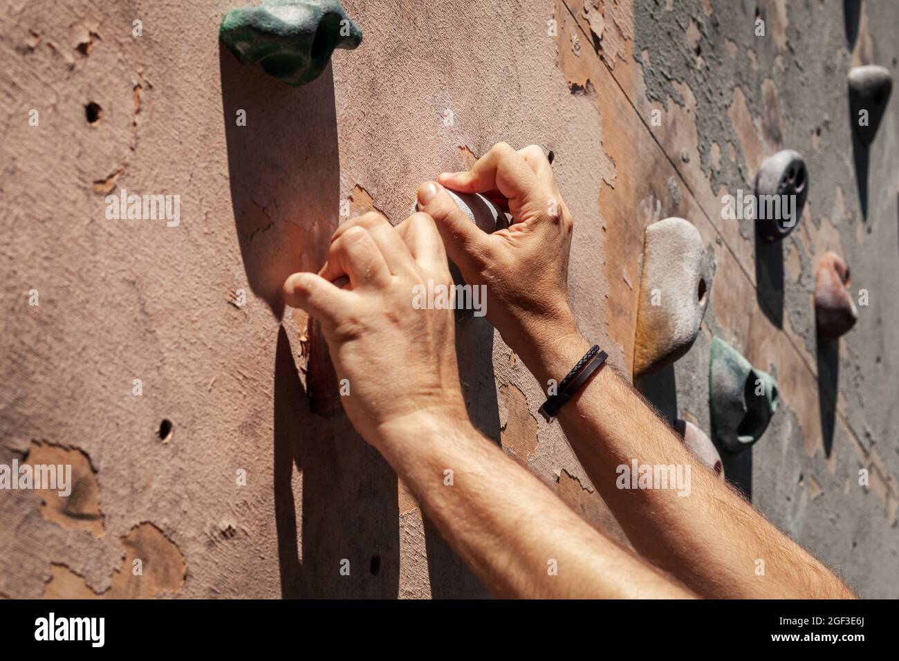 Close-up photo of male hands gripping climbing holds on worn wall outside. Man doing climbing exercise on artificial rock climbing wall at park. Stock Photo