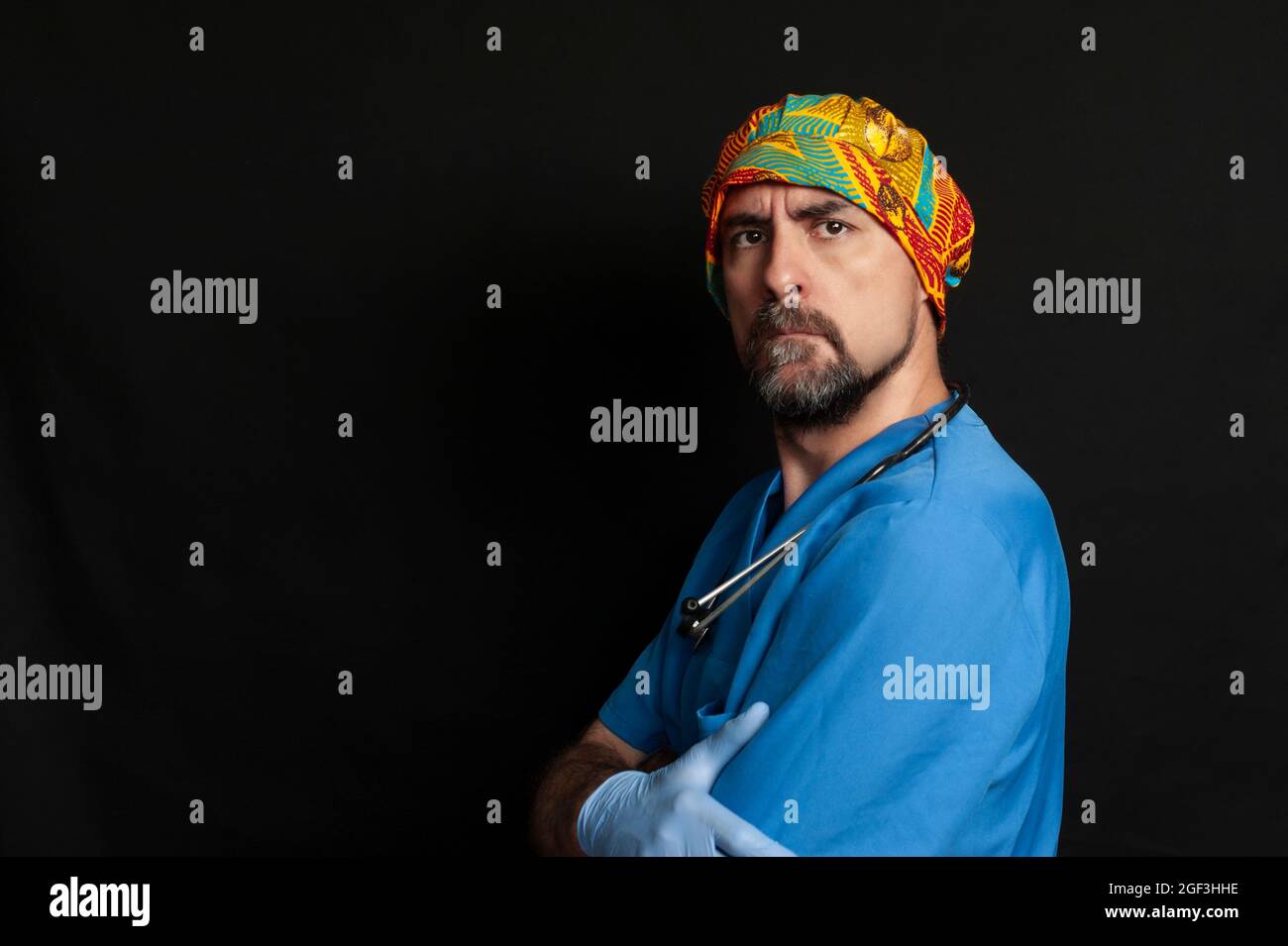 Doctor on the side looking straight ahead with a serious look on his face and his arms crossed. He is wearing a blue scrub shirt, blue surgical gloves Stock Photo