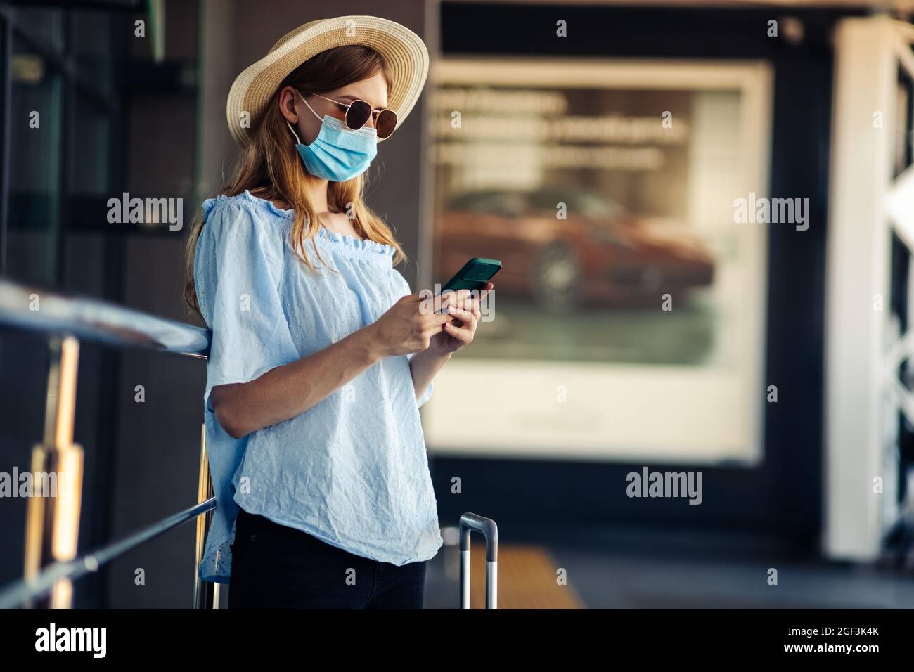Young traveler in a medical protective mask on her face, a woman walks with suitcases and uses a mobile phone, over the airport building, Concept of t Stock Photo