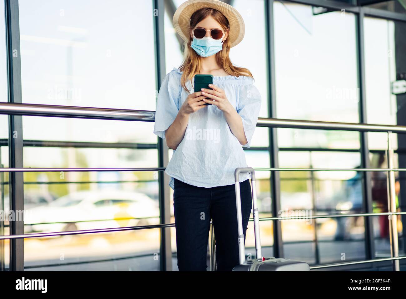 Young traveler in a medical protective mask on her face, a woman walks with suitcases and uses a mobile phone, over the airport building, Concept of t Stock Photo