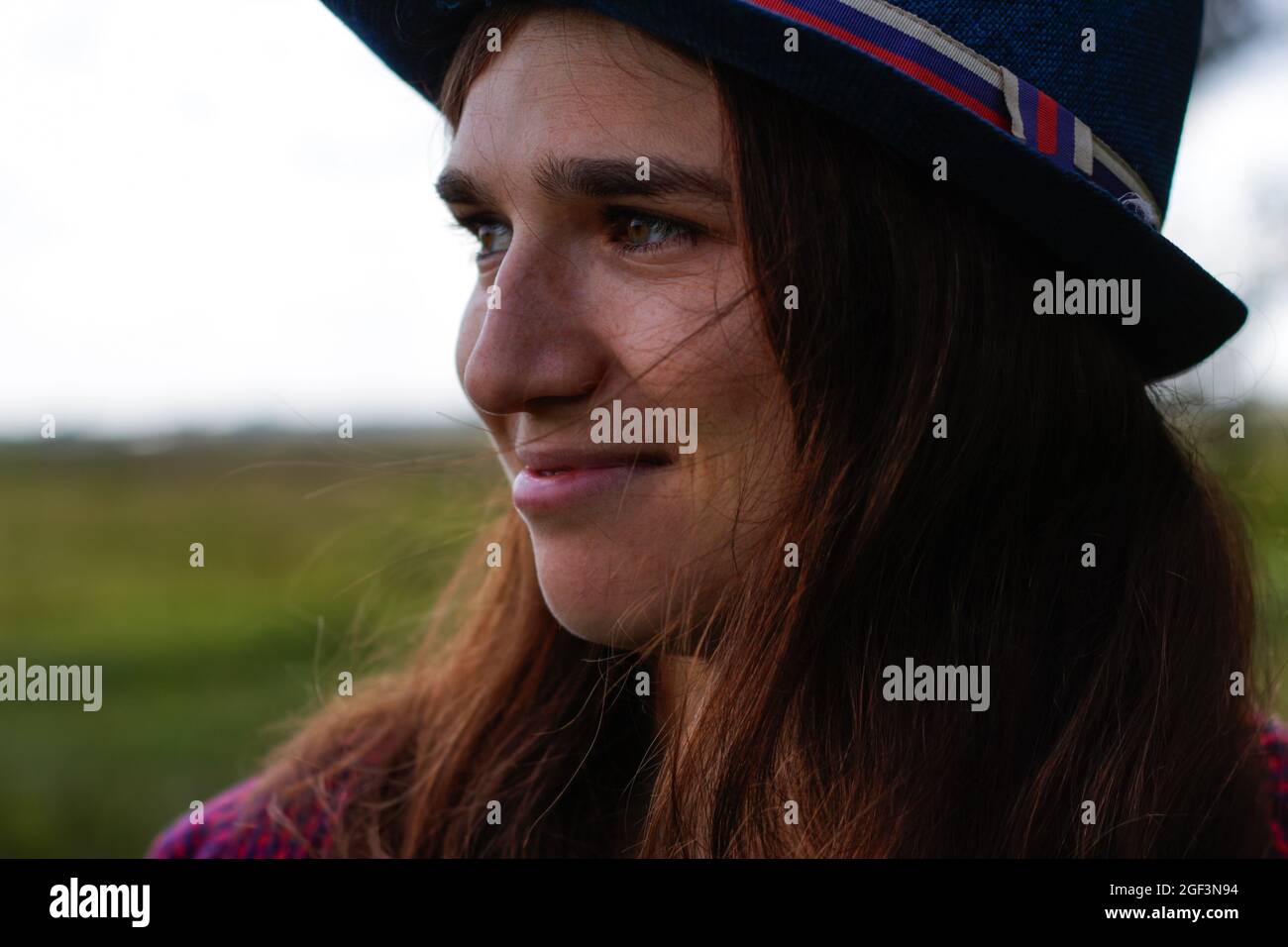 Defocus close-up portrait of a happy young woman with brown hair wearing a hat outdoors. Green nature background. Looking away. Out of focus Stock Photo