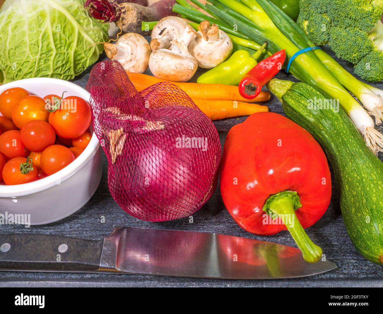 Closeup POV shot of a range of a range of raw vegetables, behind a steel chopping knife on a board, ready to cut. Stock Photo