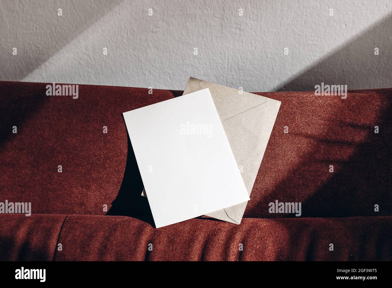 Blank greeting card mockup, envelope on rusty velvet sofa in sunlight. Empty invitation over white wall with shadows. Detail of elegant interior Stock Photo