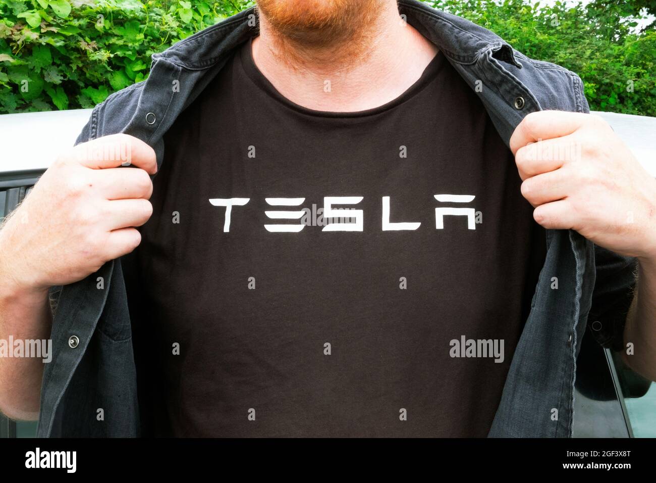 Front view of young man who admires Elon Musk energy companies holding open shirt with hands to reveal TESLA logo on t shirt in Wales UK  KATHY DEWITT Stock Photo