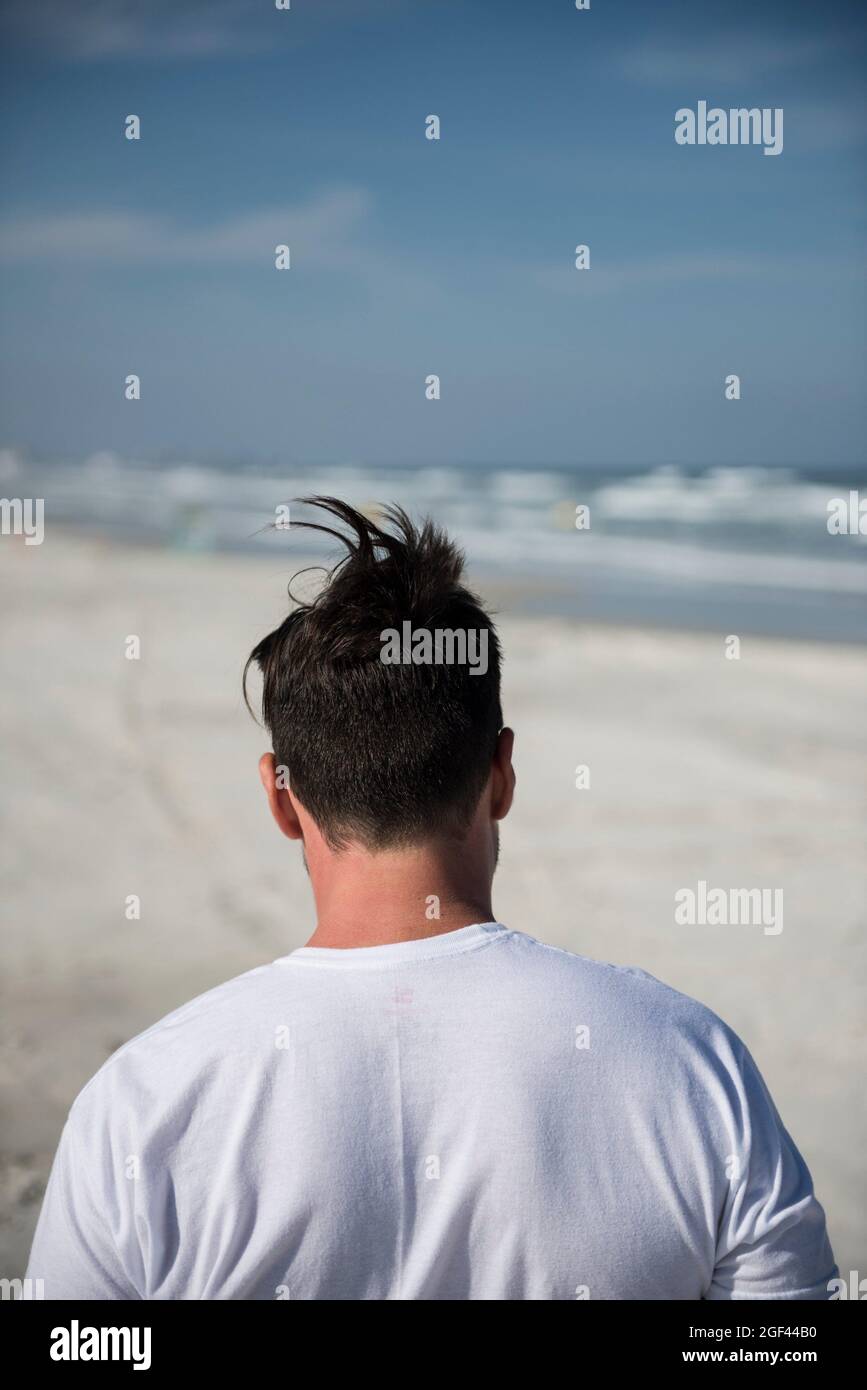 Close-up of head, neck and shoulders of a man seen from behind walking on the beach with wind blowing hair into a humorous point on top of his head. Stock Photo