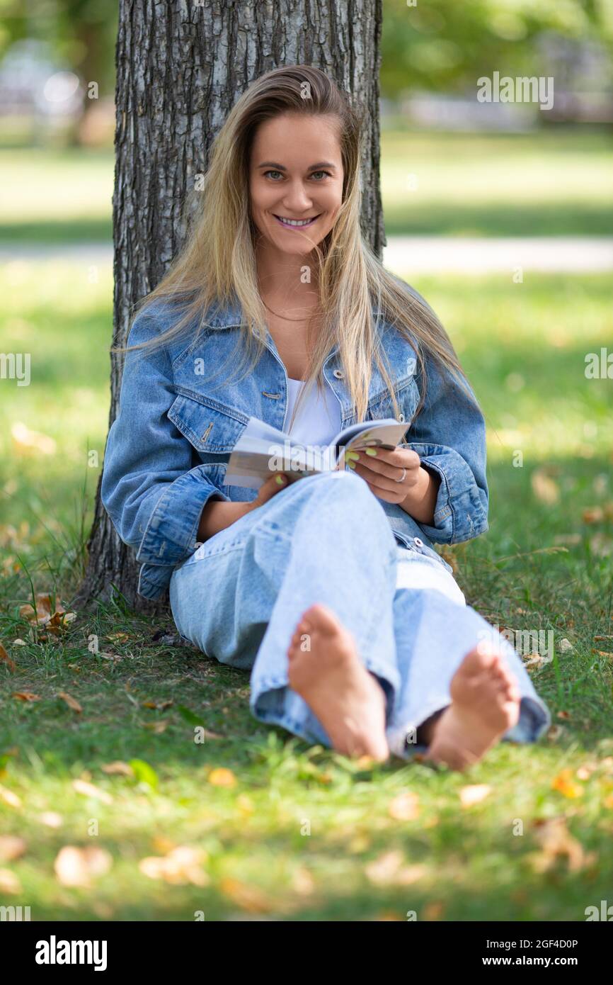 Portrait of a woman reading a book sitting in the park on the grass. She smiles and looking to the camera. The concept of rest from gadgets and city b Stock Photo