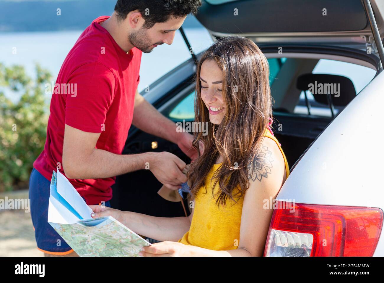 Smiling woman checking map while sitting in car trunk by boyfriend Stock Photo