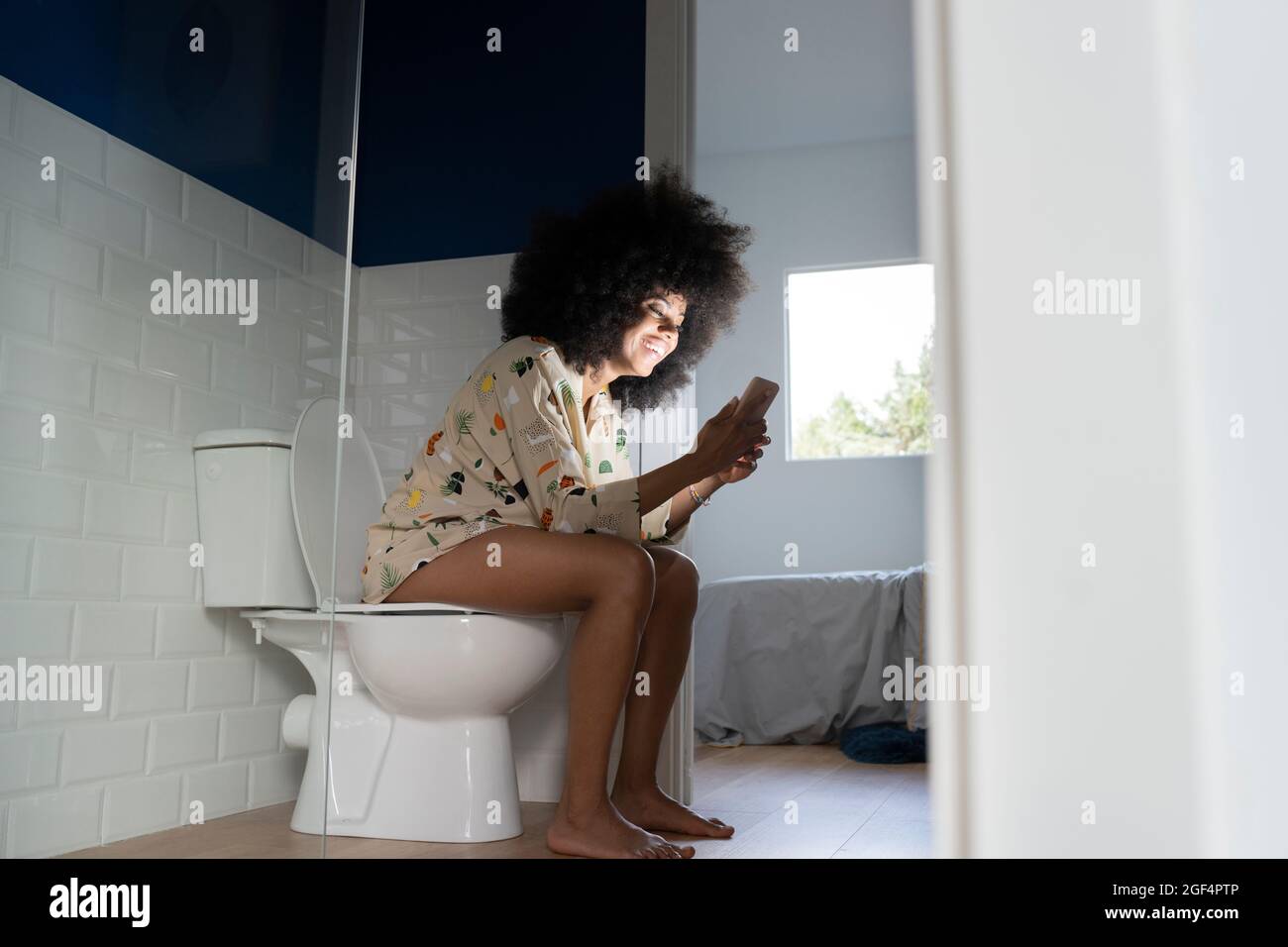 Smiling woman using smart phone while sitting on toilet seat at home Stock Photo