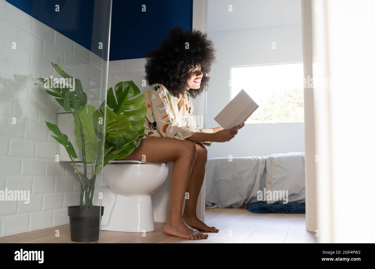 Afro woman reading book near potted plant while sitting on toilet seat at home Stock Photo
