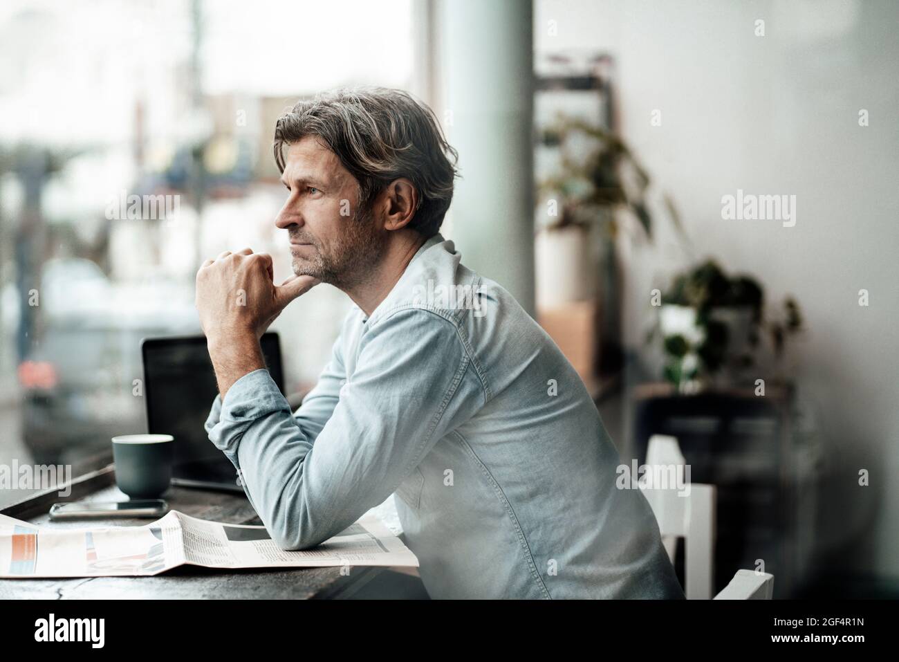 Thoughtful man sitting with newspaper looking through window while sitting in cafe Stock Photo