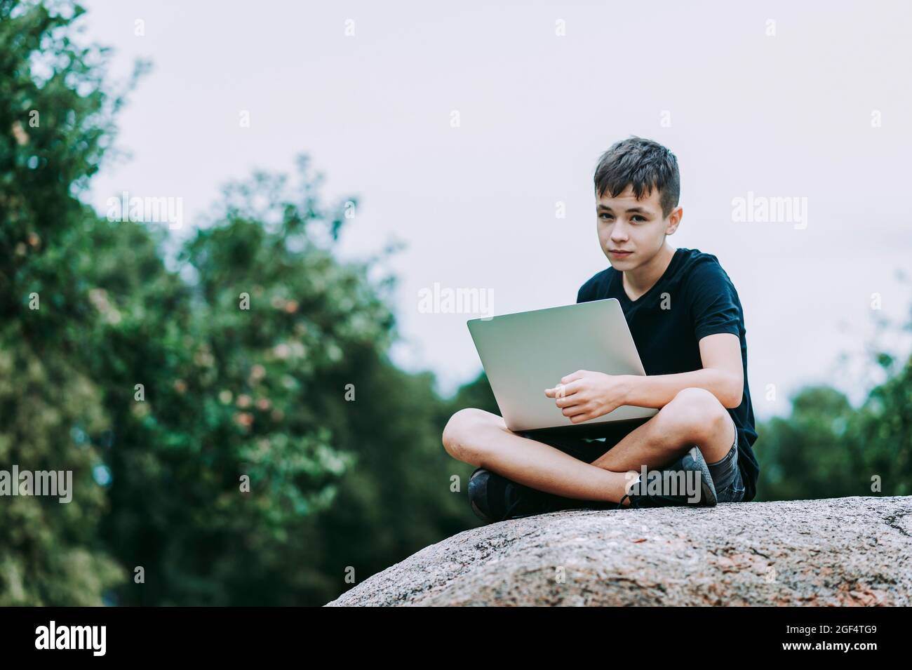 Boy sitting on a stone with laptop in garden. Kid have online web lesson or class on computer at nature, green class. Back to school. Outdoor learning Stock Photo
