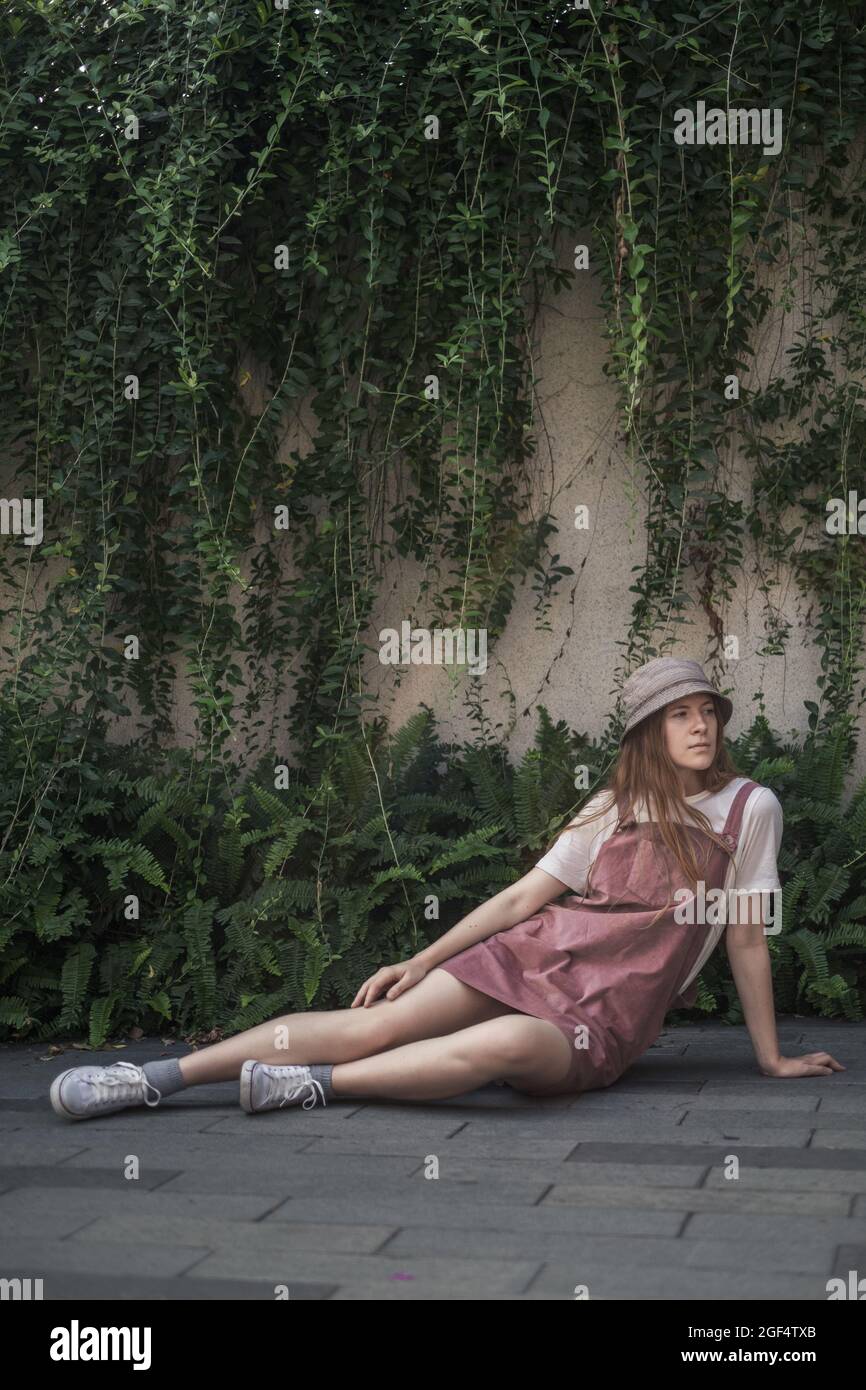 Pretty young woman in white t-shirt, pink sundress and sneakers sitting on the street. Trendy casual summer or spring outfit with the hat.  Stock Photo