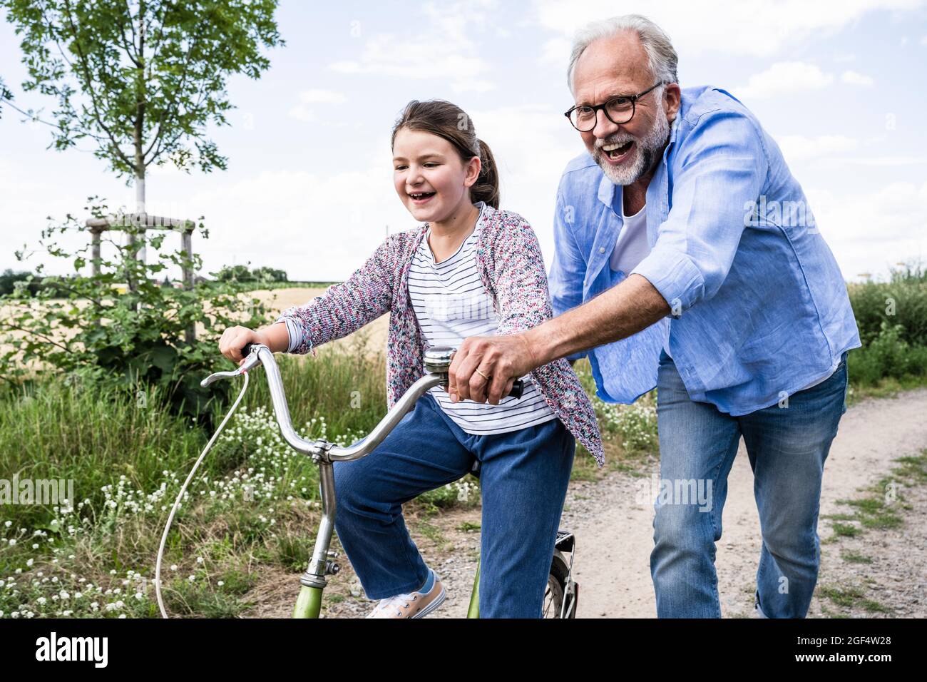 Cheerful grandfather playing with granddaughter cycling on dirt road Stock Photo