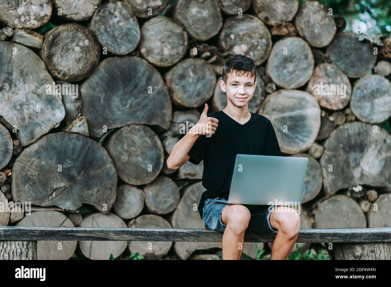 Boy sitting on wooden seat with laptop in forest. Kid have online web lesson or class on computer at nature, green class. Back to school. Outdoor lear Stock Photo