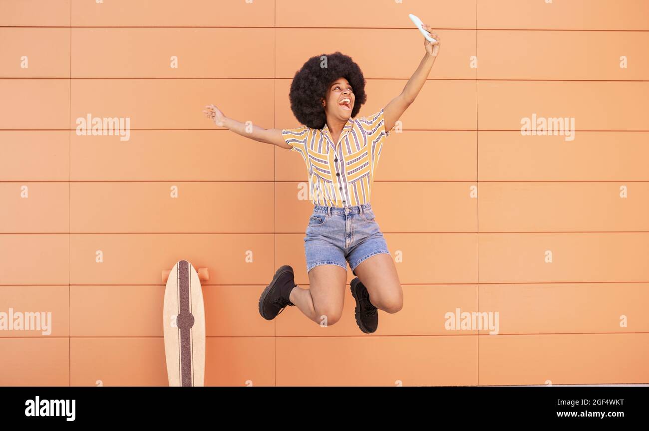 Happy woman taking selfie through smart phone while jumping in front of wall Stock Photo