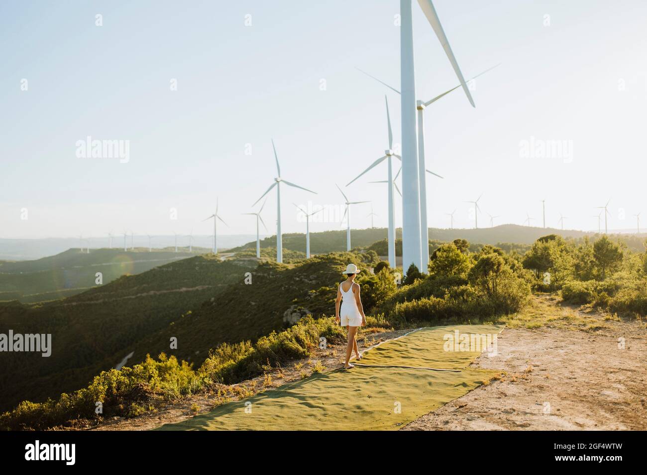 Woman walking on mountain path towards wind turbines during sunset Stock Photo