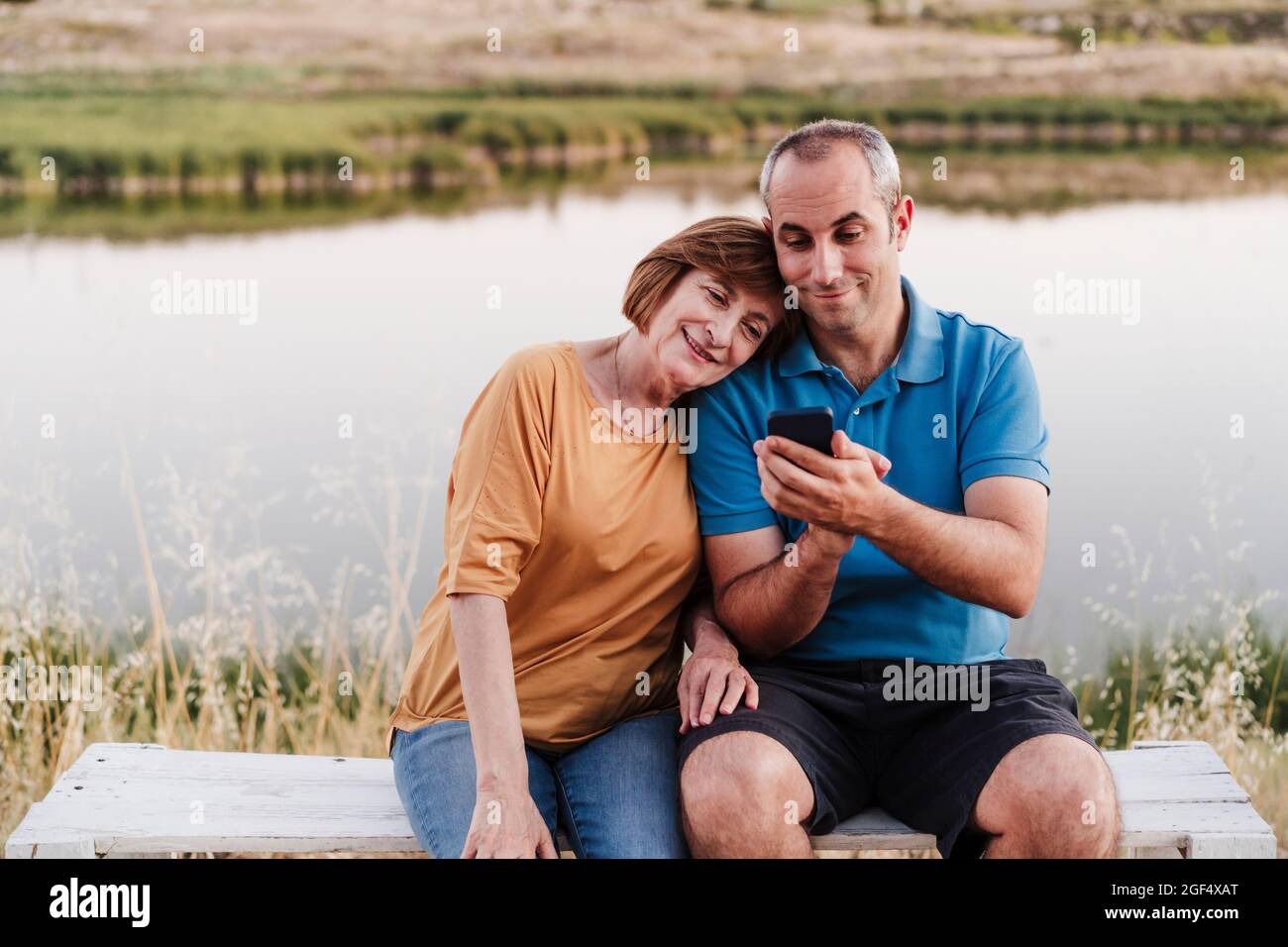 Son using smart phone while sitting with mother on bench near lake ...