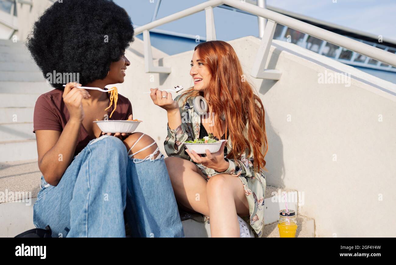 Female friends eating while sitting on steps Stock Photo