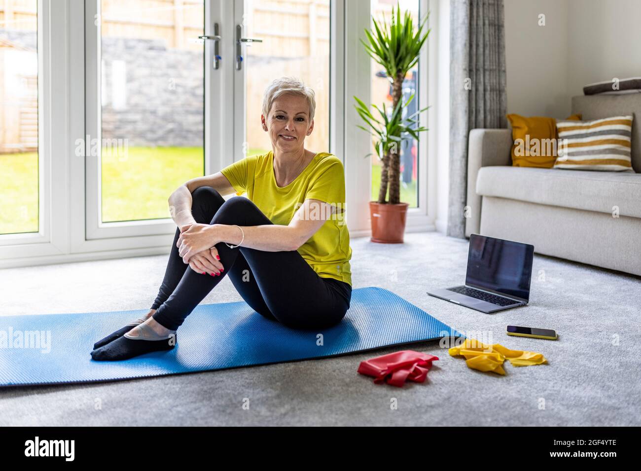 Mature woman sitting on exercise mat at home Stock Photo