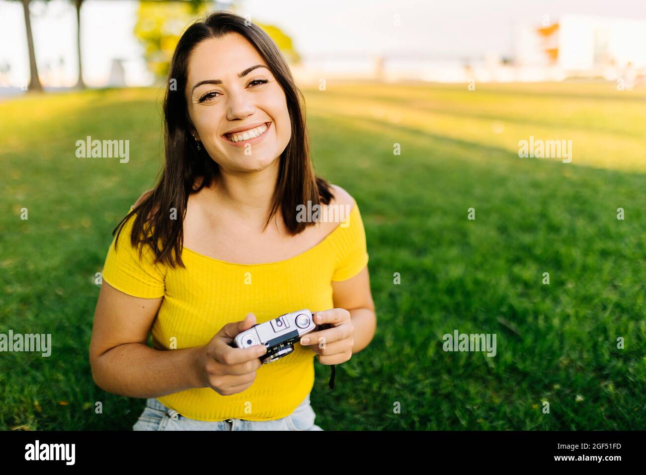 Happy young woman in off shoulder top holding camera at lawn Stock Photo