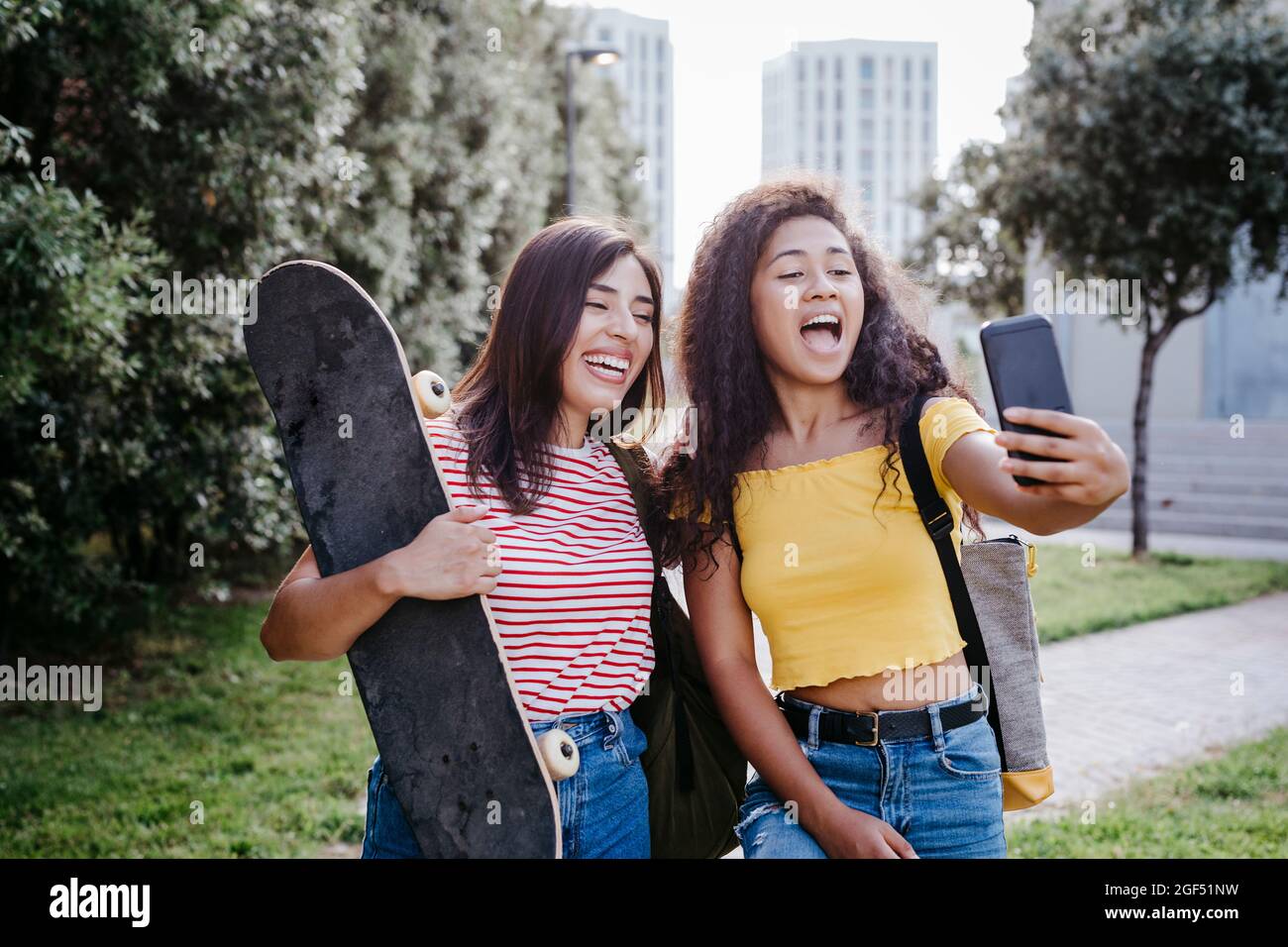 Young woman taking selfie through smart phone with female friend holding skateboard at park Stock Photo