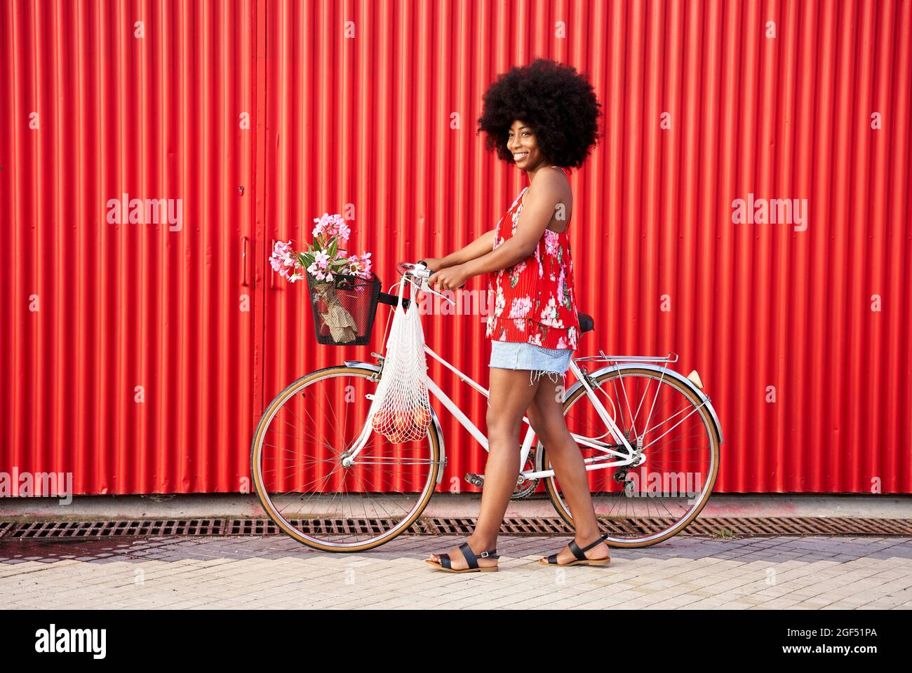 Smiling woman walking with bicycle by red corrugated wall Stock Photo