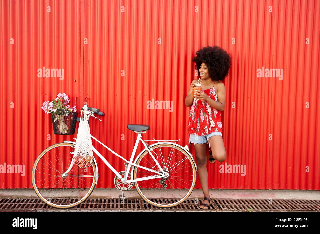 Smiling woman holding smoothie while standing by bicycle in front of red wall Stock Photo