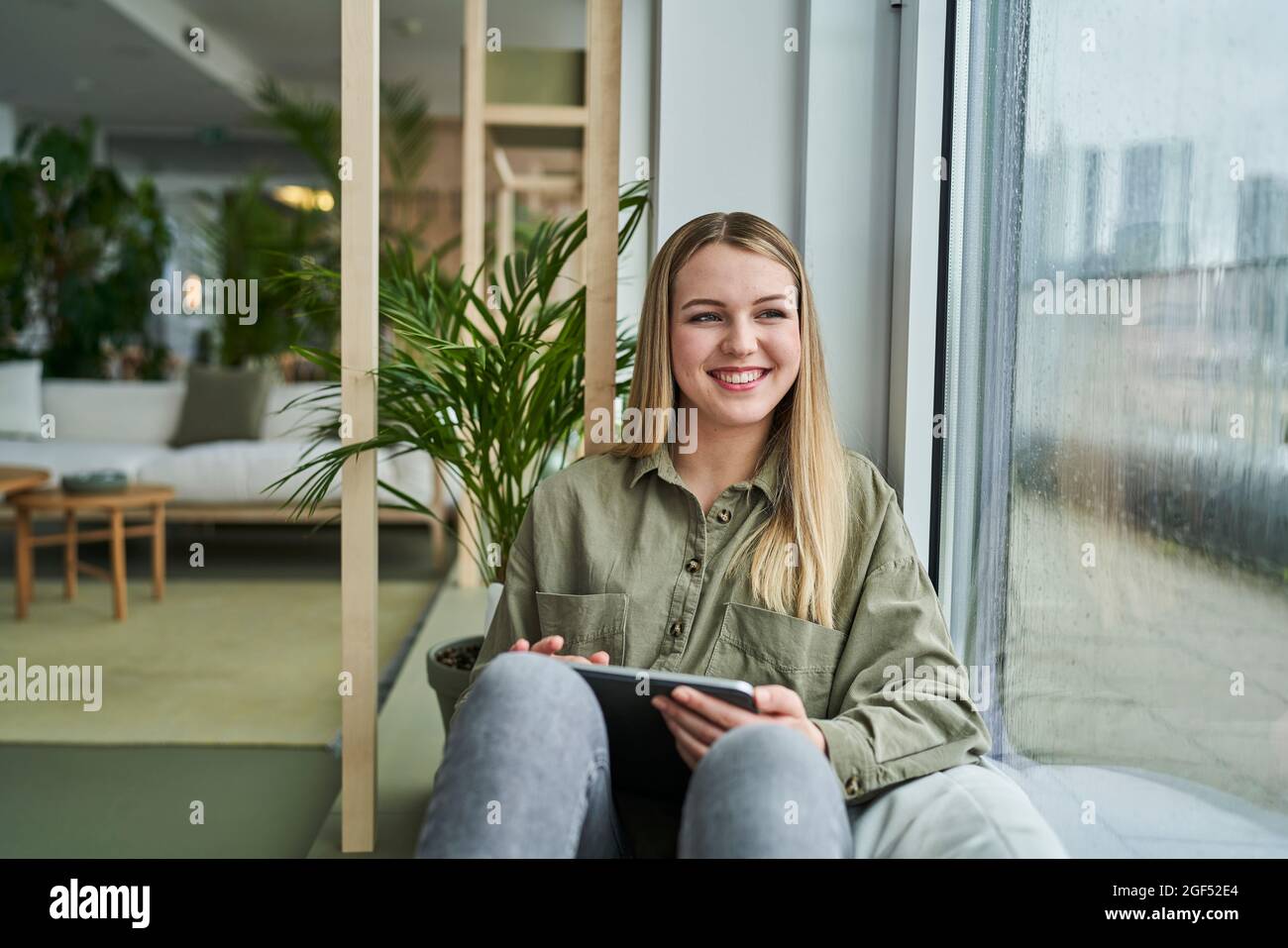 Smiling teenage female intern with digital tablet looking through window while sitting in office Stock Photo