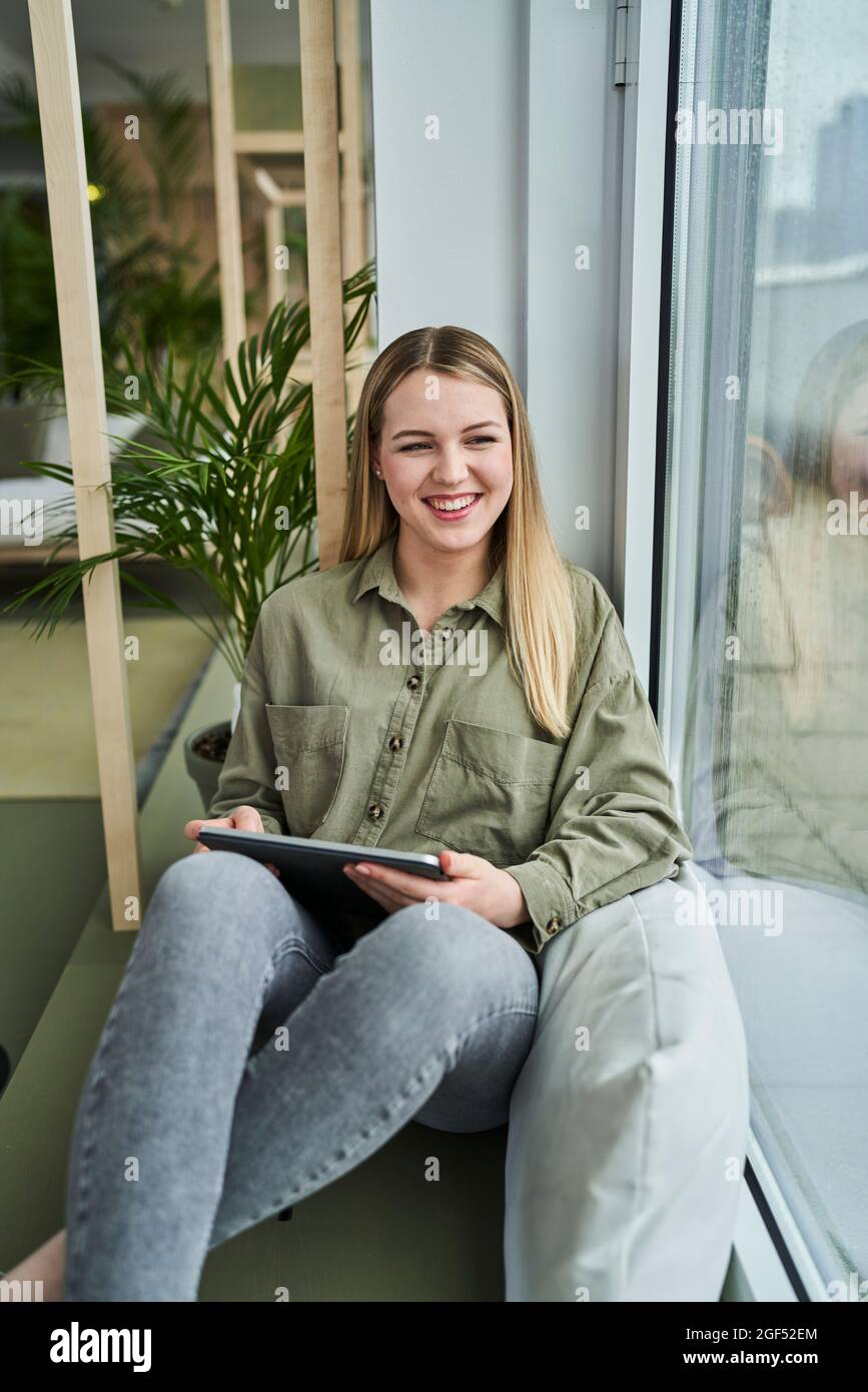 Happy female intern sitting with digital tablet by glass window at office Stock Photo