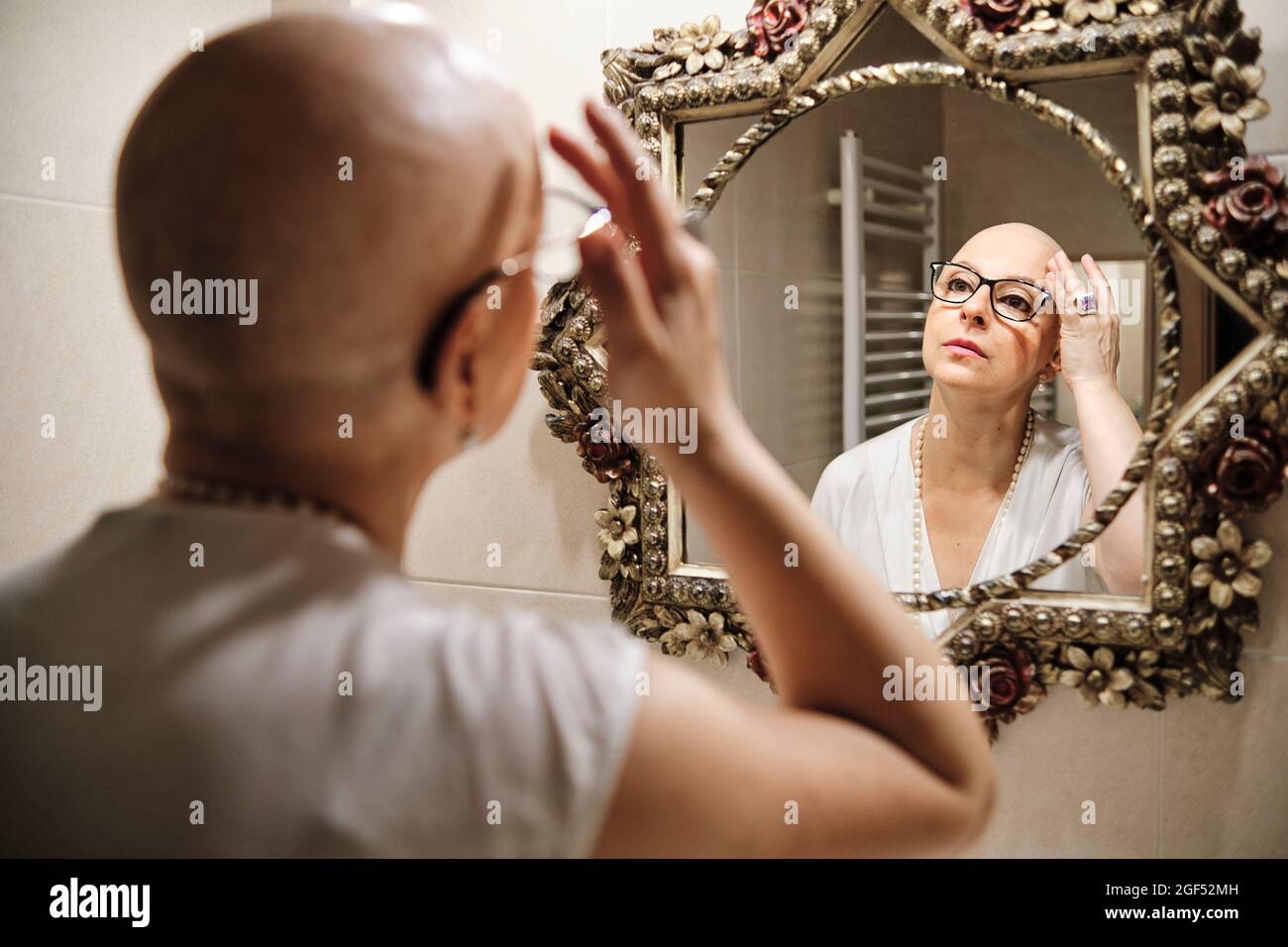 Woman with cancer touching bald head while looking at reflection in mirror Stock Photo