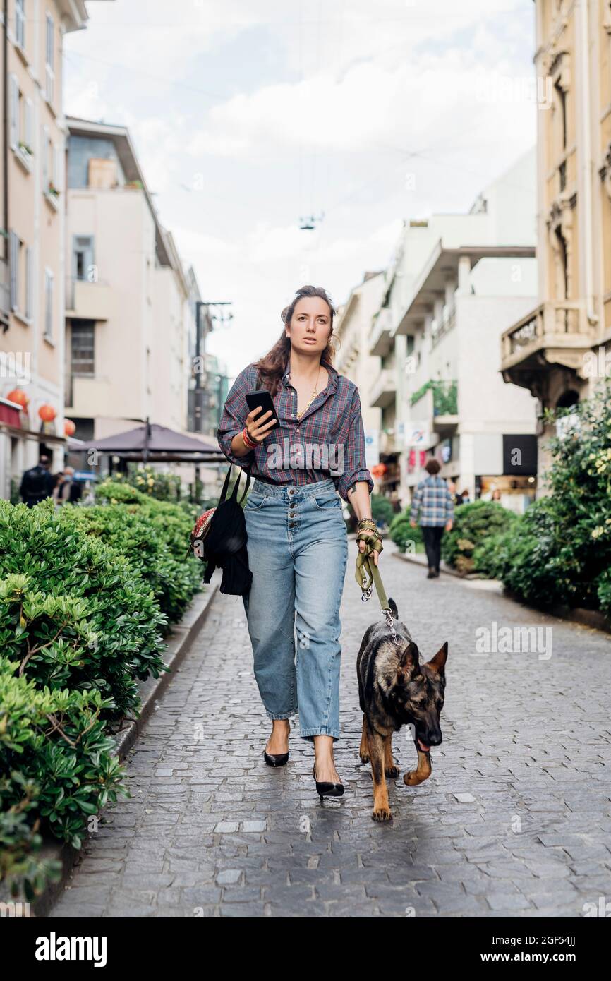 Woman holding mobile phone while walking with dog on footpath Stock Photo